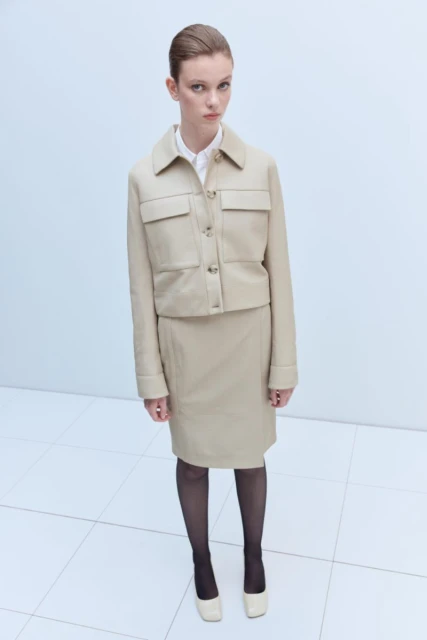 A fashionable adult woman is posing in a beige jacket and skirt outfit with black stockings and matching shoes on a white tile floor.