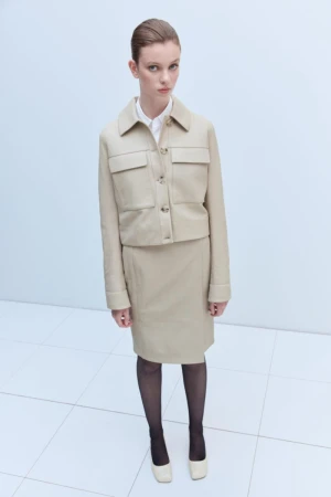 A fashionable adult woman is posing in a beige jacket and skirt outfit with black stockings and matching shoes on a white tile floor.
