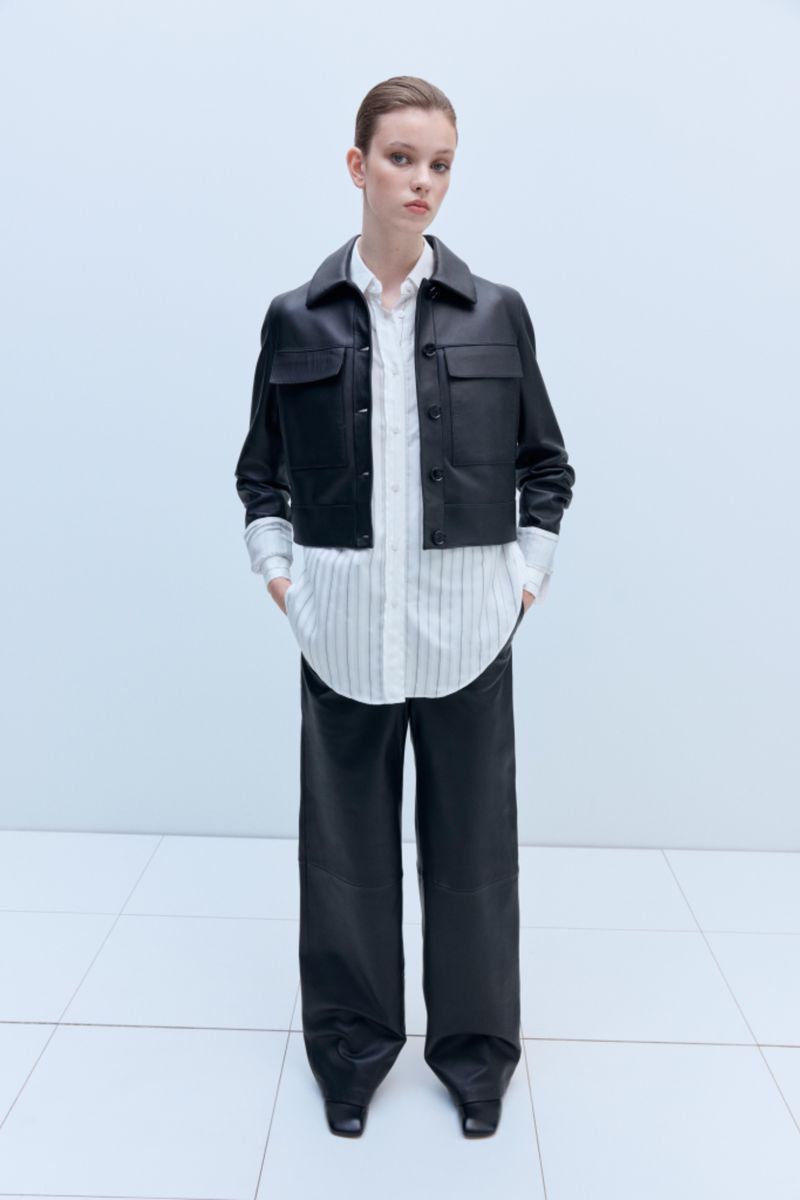 Fashionable woman in a black leather jacket and wide-leg pants stands against a light background in a studio. Full-length fashion shot.