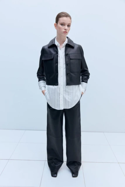 Fashionable woman in a black leather jacket and wide-leg pants stands against a light background in a studio. Full-length fashion shot.