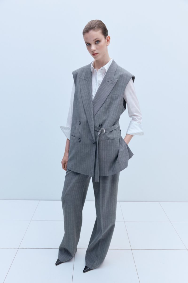 Woman in grey pinstripe vest suit and white shirt standing indoors in a fashion pose against a white background with square tiling.