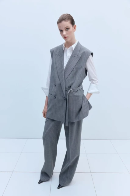 Woman in grey pinstripe vest suit and white shirt standing indoors in a fashion pose against a white background with square tiling.