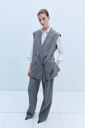 Woman in grey pinstripe vest suit and white shirt standing indoors in a fashion pose against a white background with square tiling.
