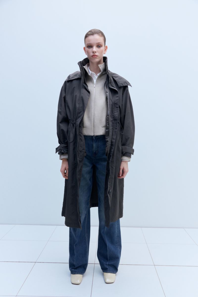 Full-length shot of a young adult woman in jeans, zip-up jacket, and long dark gray coat, posing in a neutral indoor setting with white floors.