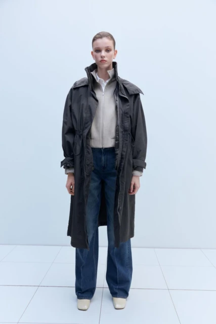 Full-length shot of a young adult woman in jeans, zip-up jacket, and long dark gray coat, posing in a neutral indoor setting with white floors.
