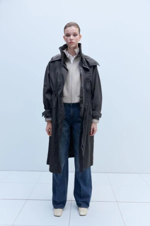 Full-length shot of a young adult woman in jeans, zip-up jacket, and long dark gray coat, posing in a neutral indoor setting with white floors.