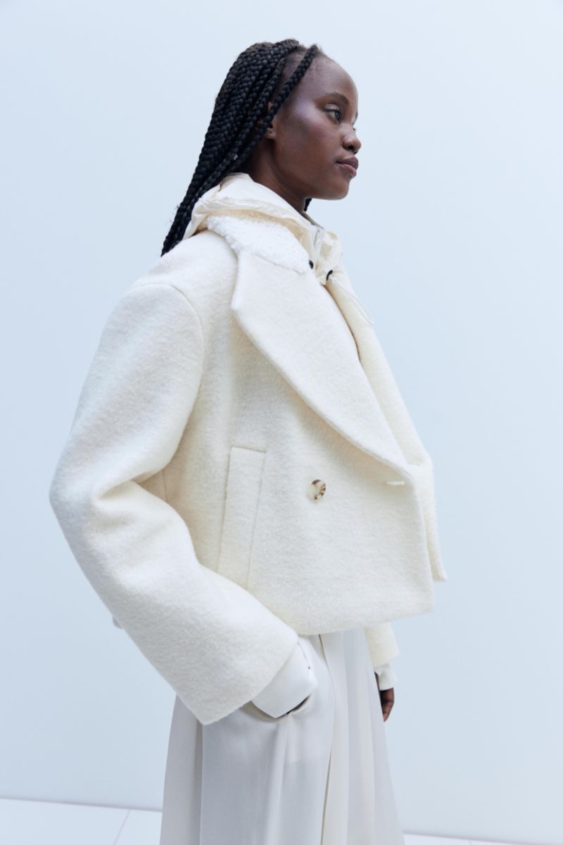 An African woman in profile wears a cream-colored wool peacoat with an oversized collar and tailored skirt in a fashion studio shot.