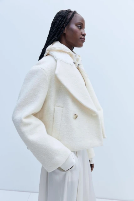An African woman in profile wears a cream-colored wool peacoat with an oversized collar and tailored skirt in a fashion studio shot.