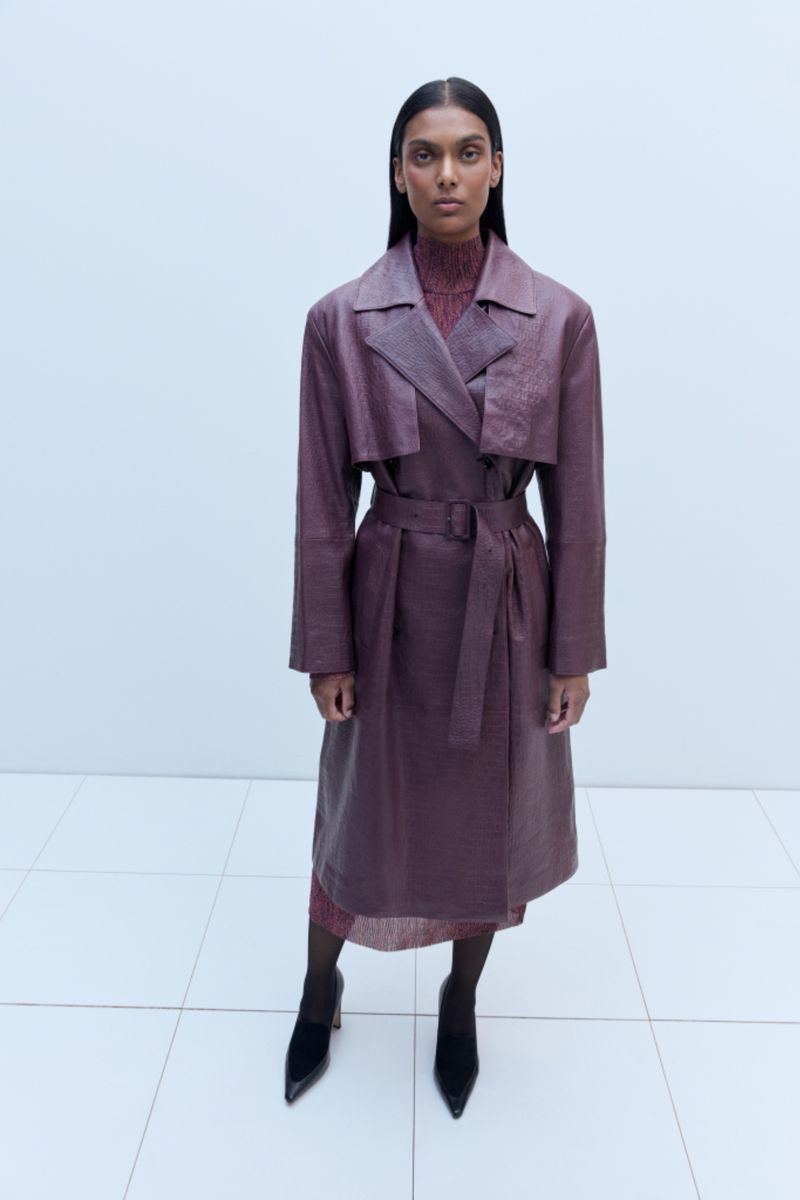 A woman in a long purple textured coat and black heels stands in a white studio, looking directly at the camera in a fashion shot.
