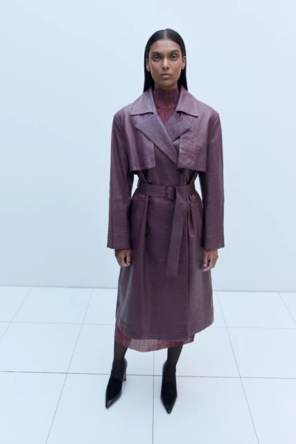 A woman in a long purple textured coat and black heels stands in a white studio, looking directly at the camera in a fashion shot.