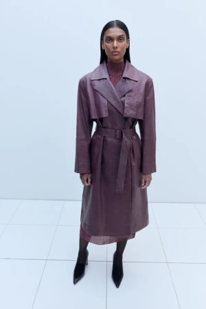 A woman in a long purple textured coat and black heels stands in a white studio, looking directly at the camera in a fashion shot.