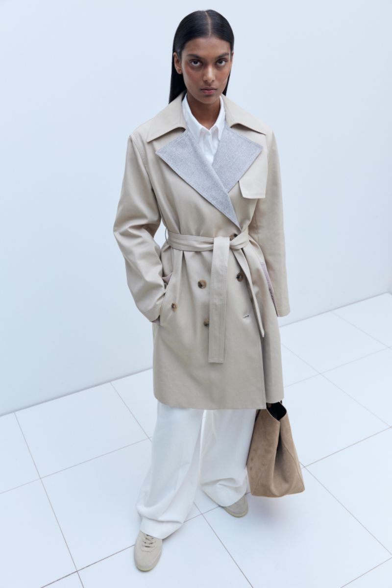 Woman in a tan trench coat, white pants, and sneakers standing indoors on a white tiled floor, carrying a tan tote bag over her shoulder.