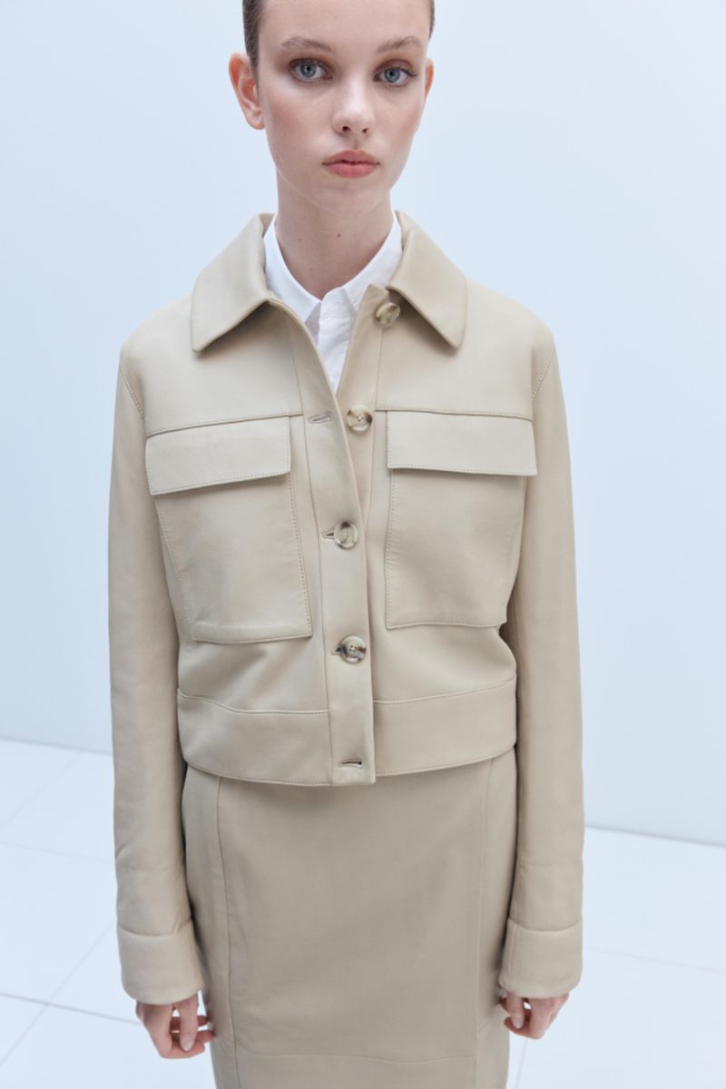 Adult female posing in a beige skirt and jacket in a studio. She wears a white collared shirt and jacket with visible buttons and pockets.