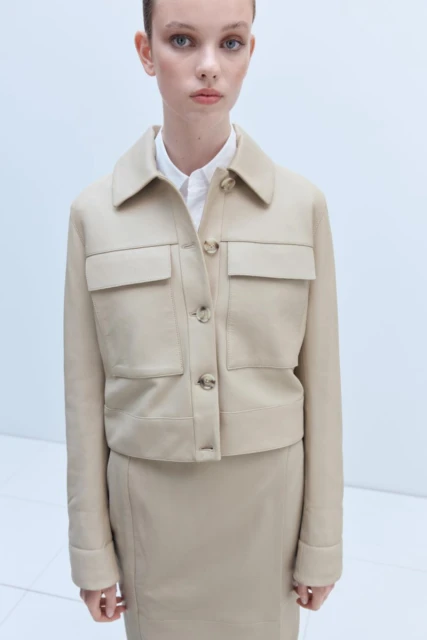 Adult female posing in a beige skirt and jacket in a studio. She wears a white collared shirt and jacket with visible buttons and pockets.