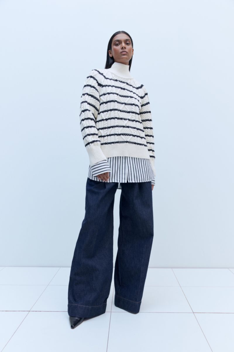 Brunette woman poses in a cream and black striped sweater, blue wide-leg jeans, and black heels in a minimalist, light-filled studio.
