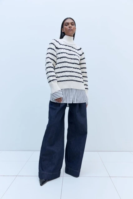 Brunette woman poses in a cream and black striped sweater, blue wide-leg jeans, and black heels in a minimalist, light-filled studio.