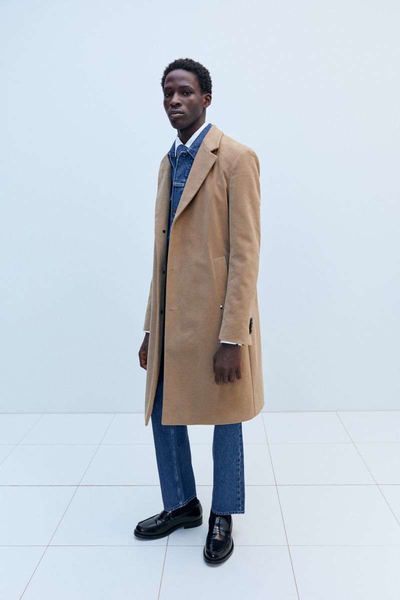 A lean, dark-skinned man in a tan overcoat, denim shirt, blue jeans, and black loafers poses in front of a white background.