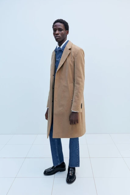 A lean, dark-skinned man in a tan overcoat, denim shirt, blue jeans, and black loafers poses in front of a white background.