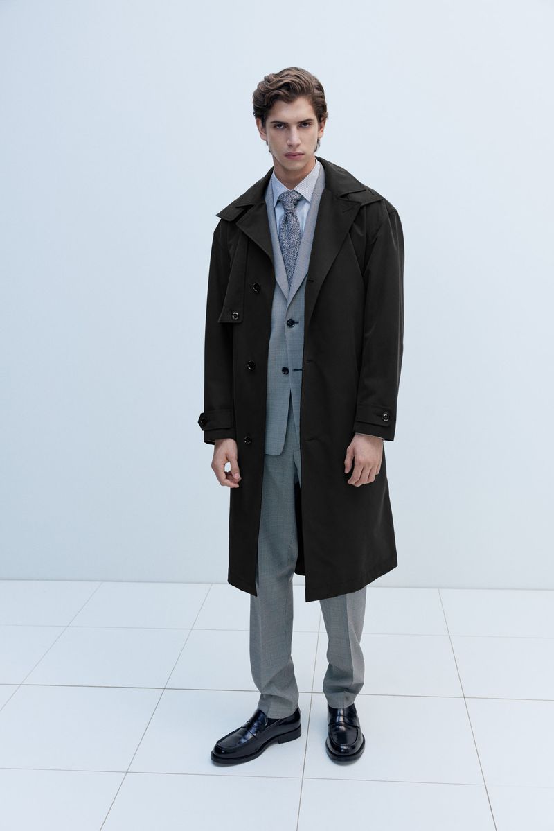 Full-length shot of an adult male in a grey suit, patterned tie, dark brown trench coat, and black dress shoes in a white studio.