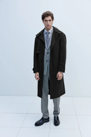 Full-length shot of an adult male in a grey suit, patterned tie, dark brown trench coat, and black dress shoes in a white studio.