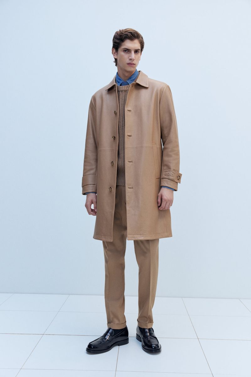 A full shot of a male model in a tan coat and matching pants stands against a bright white wall and tiled floor in a fashion editorial.