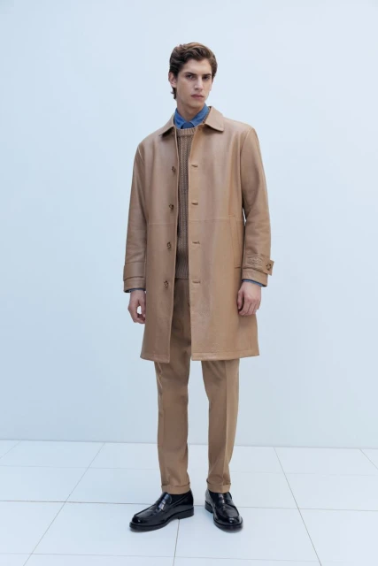 A full shot of a male model in a tan coat and matching pants stands against a bright white wall and tiled floor in a fashion editorial.
