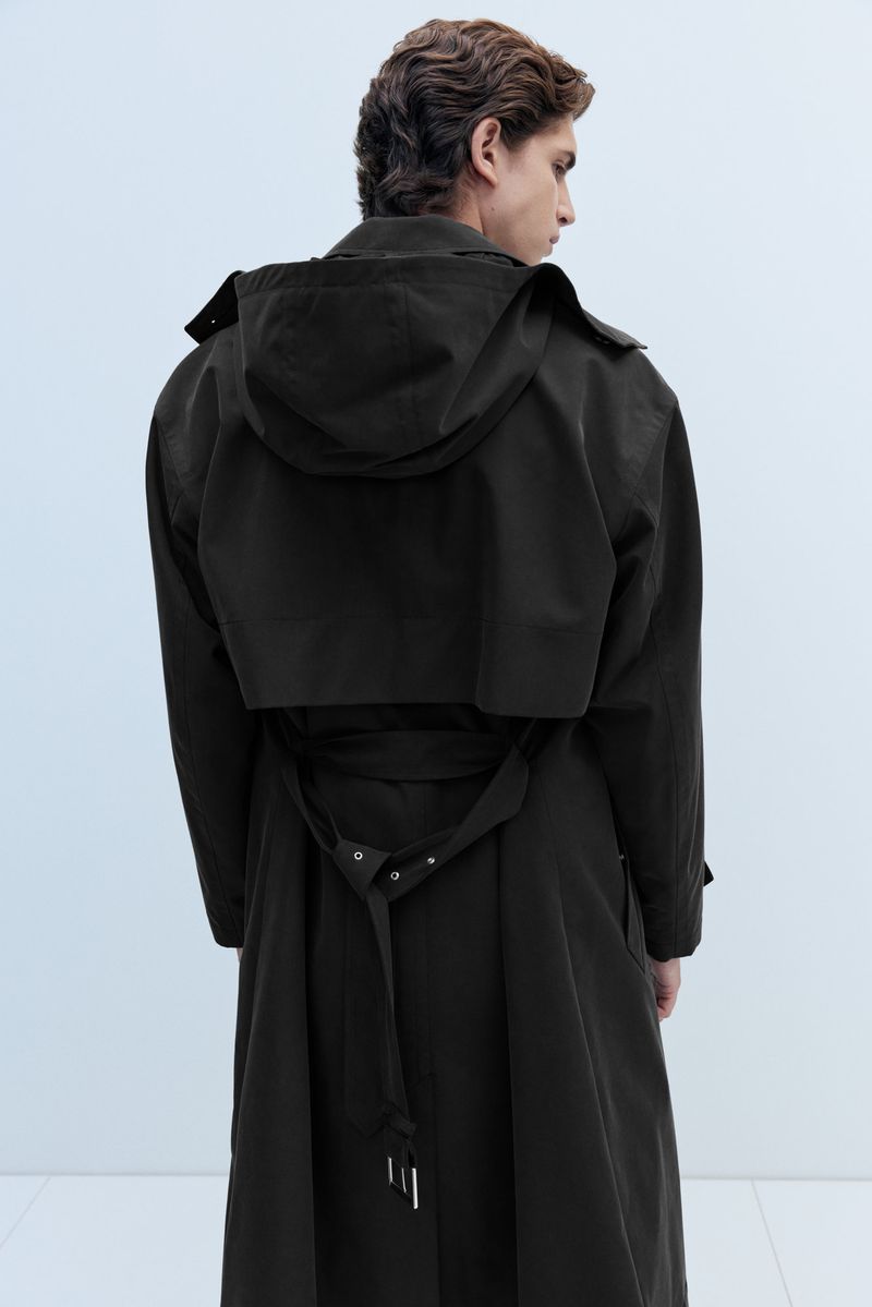 Rear view of a man wearing a hooded black belted trench coat standing against a minimalist light studio background.