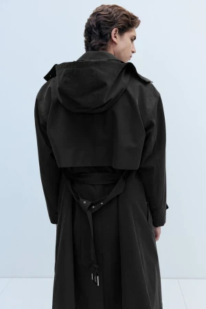 Rear view of a man wearing a hooded black belted trench coat standing against a minimalist light studio background.