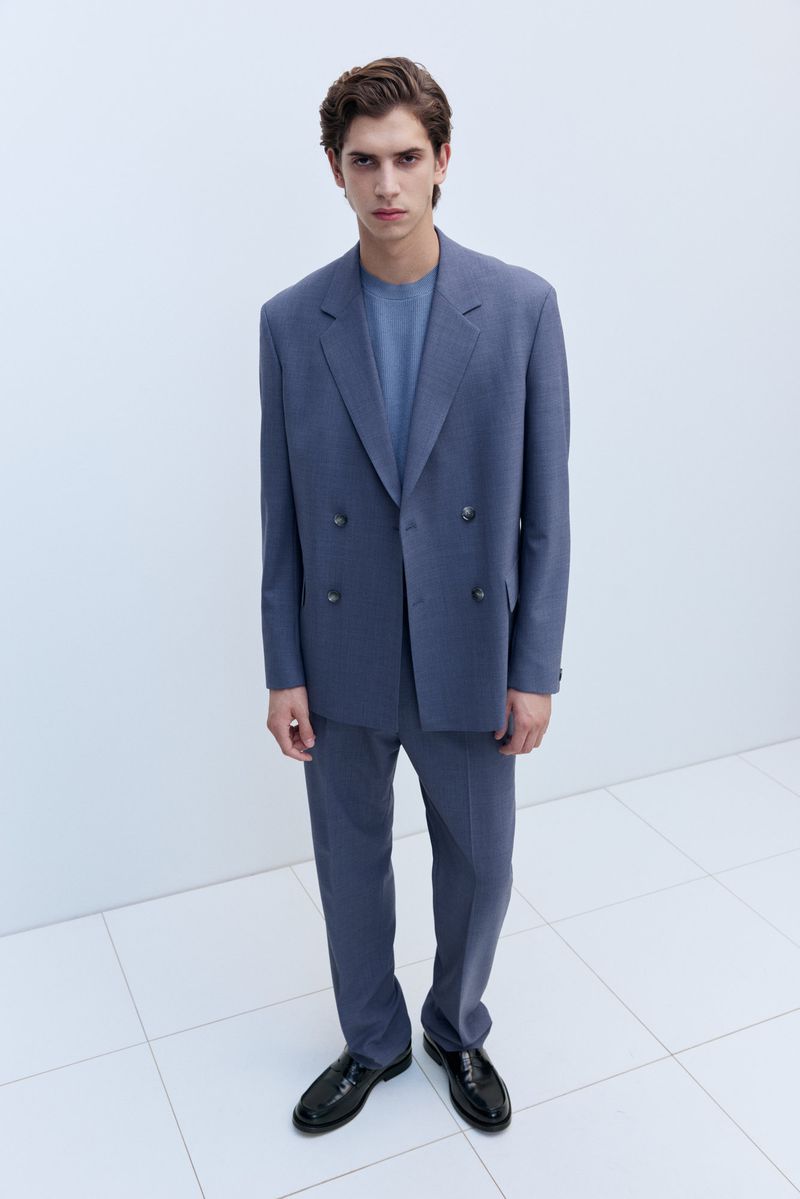 A male model poses in a blue double-breasted suit with black dress shoes on a white tiled surface, showcasing a fashion-forward formal style.