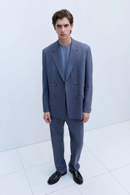 A male model poses in a blue double-breasted suit with black dress shoes on a white tiled surface, showcasing a fashion-forward formal style.