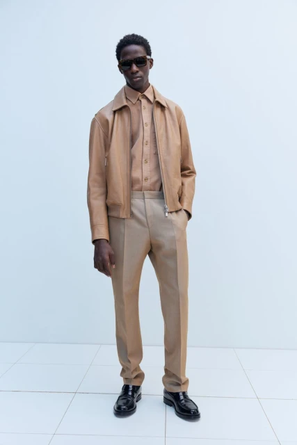Man in tan jacket, shirt, and trousers stands against a white wall wearing sunglasses and black leather shoes in full shot.