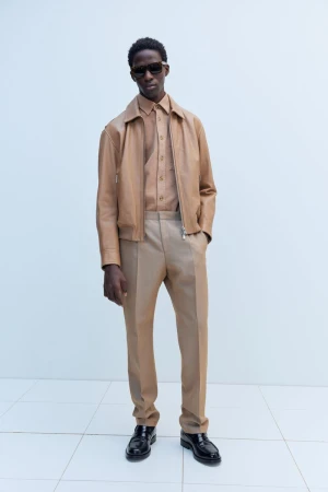 Man in tan jacket, shirt, and trousers stands against a white wall wearing sunglasses and black leather shoes in full shot.