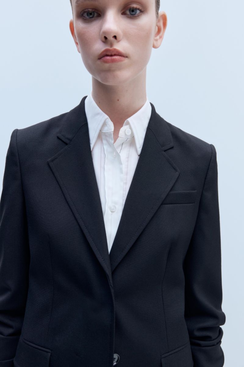 Close-up portrait of a woman wearing a tailored black blazer over a crisp white collared shirt against a pale studio backdrop.