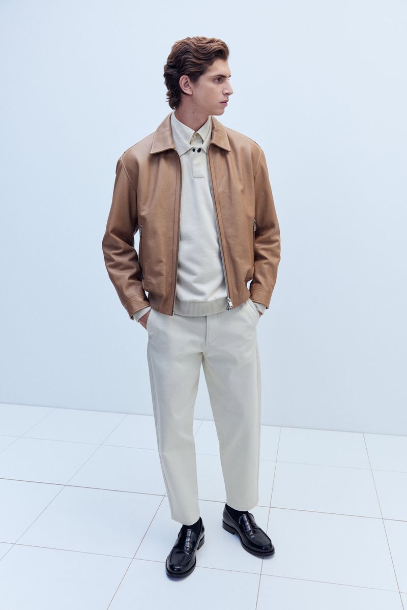 A fashionable adult male is posing in a studio wearing a brown leather jacket, a beige polo shirt, and beige pants with shiny black shoes.