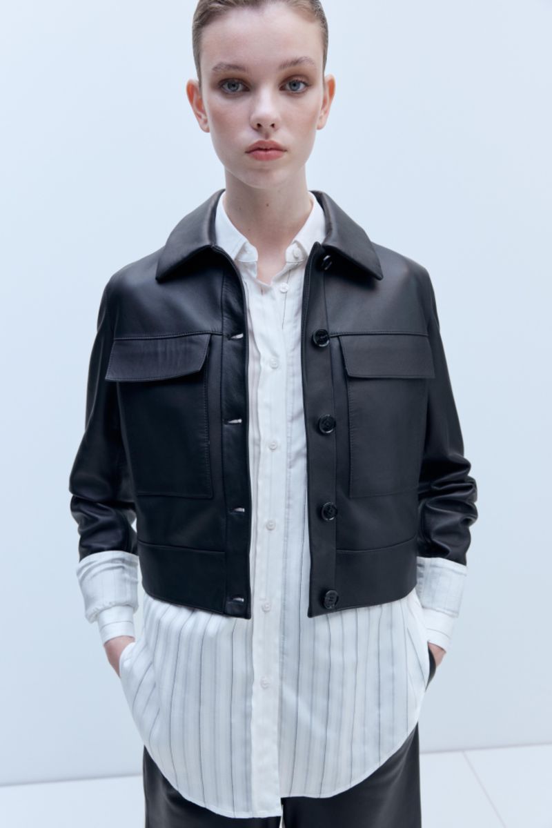 Model wearing a cropped black leather jacket layered over a long white pinstripe button-down shirt against a pale studio backdrop.