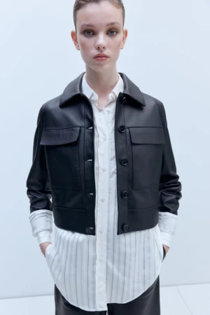 Model wearing a cropped black leather jacket layered over a long white pinstripe button-down shirt against a pale studio backdrop.