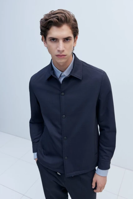 Portrait of a young adult man wearing a navy button-down jacket over a light-blue shirt, posing indoors with a neutral facial expression.