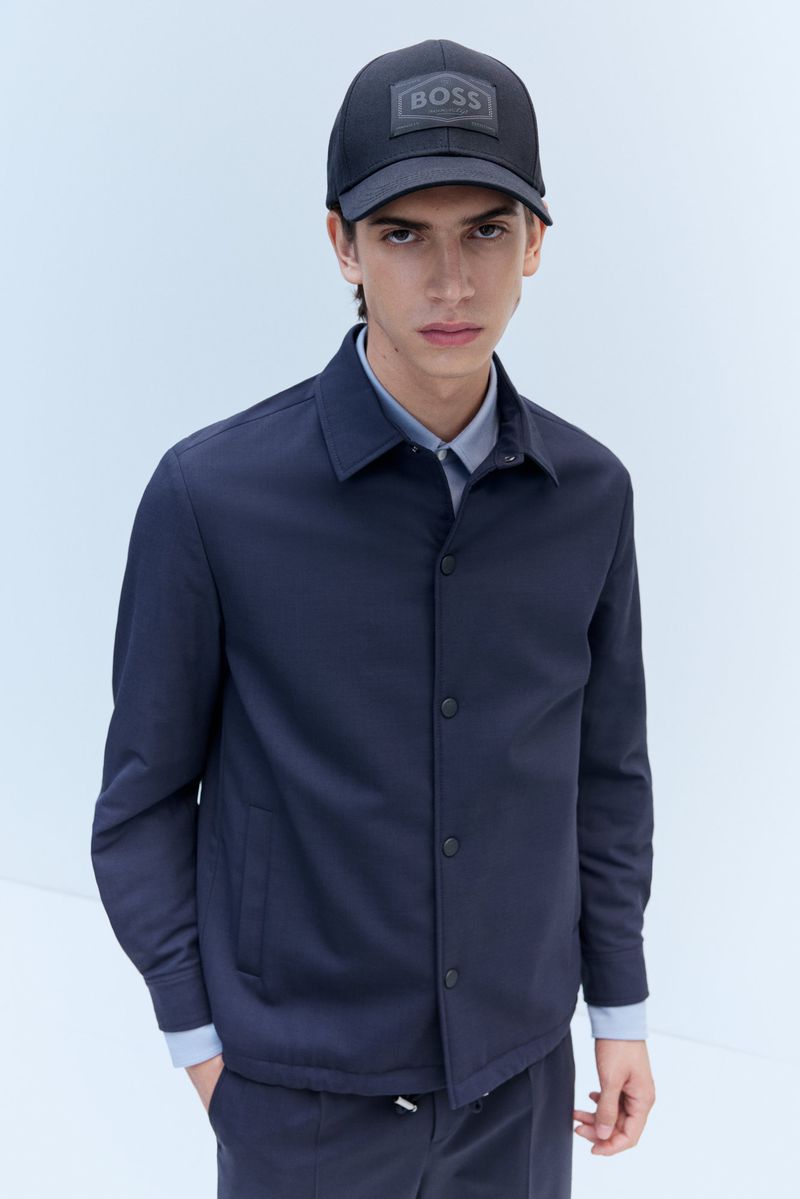 Young man in navy jacket and baseball cap with BOSS logo in studio setting. Minimalist fashion look. Medium shot on white background.