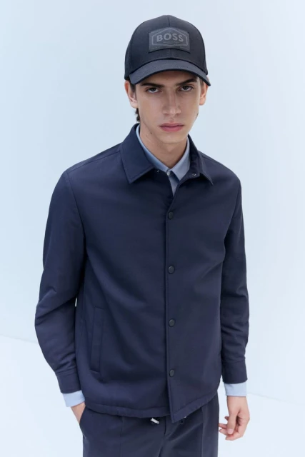 Young man in navy jacket and baseball cap with BOSS logo in studio setting. Minimalist fashion look. Medium shot on white background.
