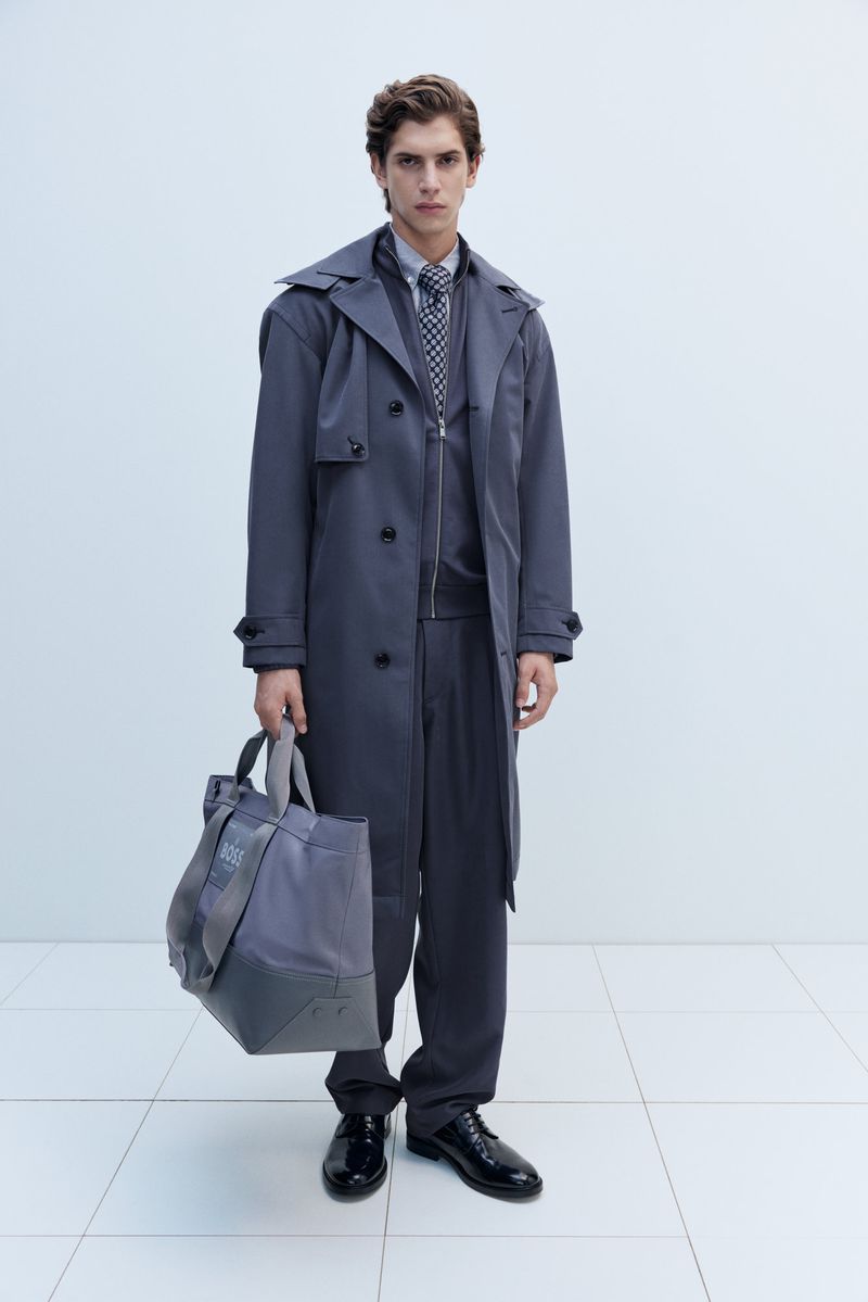 Man in grey trench coat holds tote bag in a studio shot, wearing matching suit and tie, standing against a white backdrop.