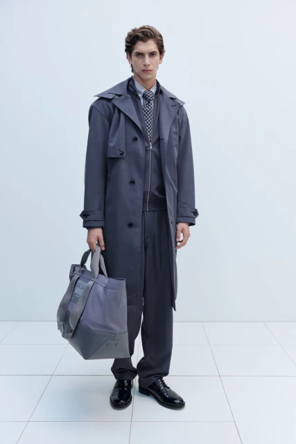 Man in grey trench coat holds tote bag in a studio shot, wearing matching suit and tie, standing against a white backdrop.