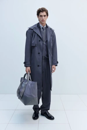 Man in grey trench coat holds tote bag in a studio shot, wearing matching suit and tie, standing against a white backdrop.