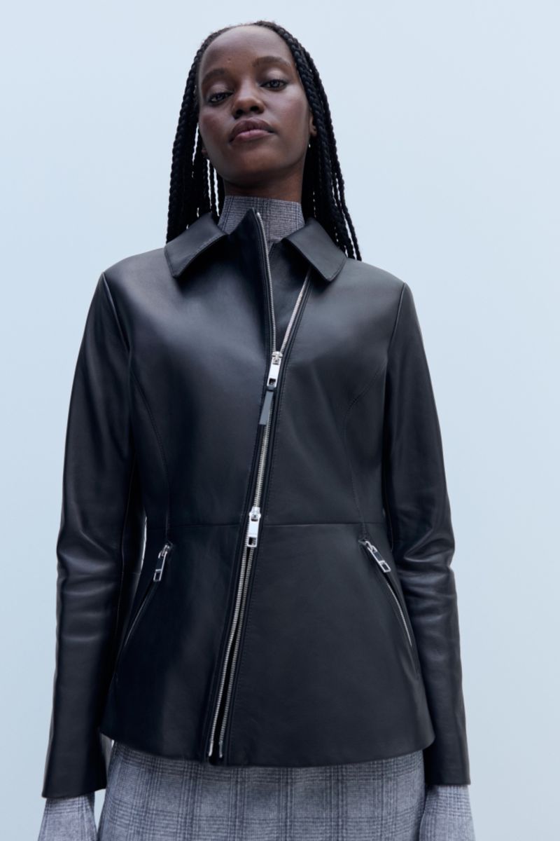 Woman wearing a black leather jacket with an asymmetrical silver zipper and a grey patterned skirt in a studio setting.