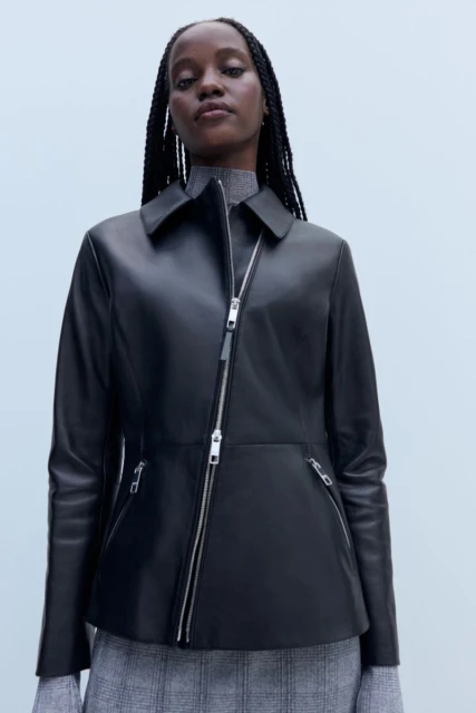 Woman wearing a black leather jacket with an asymmetrical silver zipper and a grey patterned skirt in a studio setting.