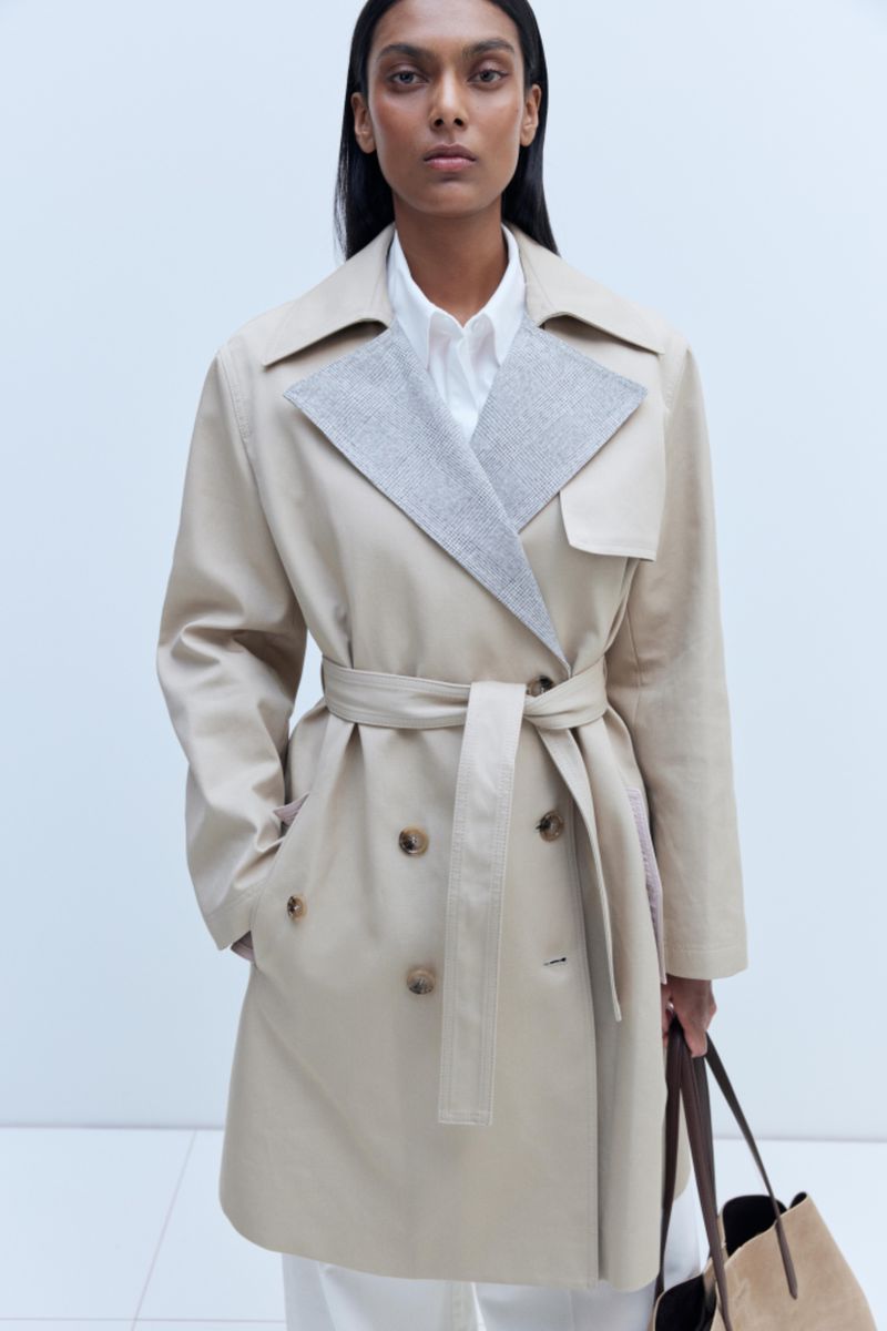 A woman models a beige trench coat with a gray collar and matching belt. She carries a tote bag against a neutral background.