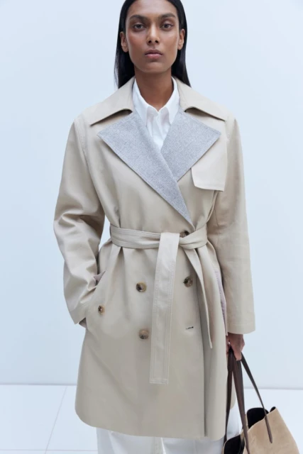 A woman models a beige trench coat with a gray collar and matching belt. She carries a tote bag against a neutral background.