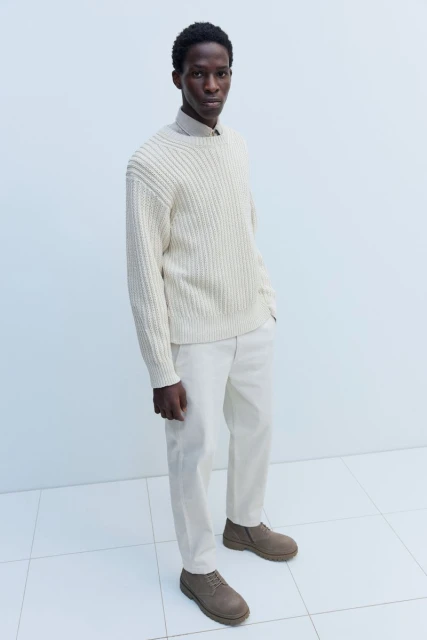 Full-length studio shot of a stylish Black man in a cream-colored knit sweater and tan trousers, posing against a pale blue background.
