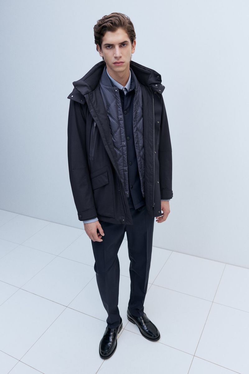 Full-length shot of a male model in a dark hooded coat, button-down shirt, dress pants and shiny leather shoes, captured from a high angle.