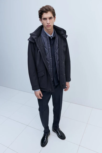 Full-length shot of a male model in a dark hooded coat, button-down shirt, dress pants and shiny leather shoes, captured from a high angle.