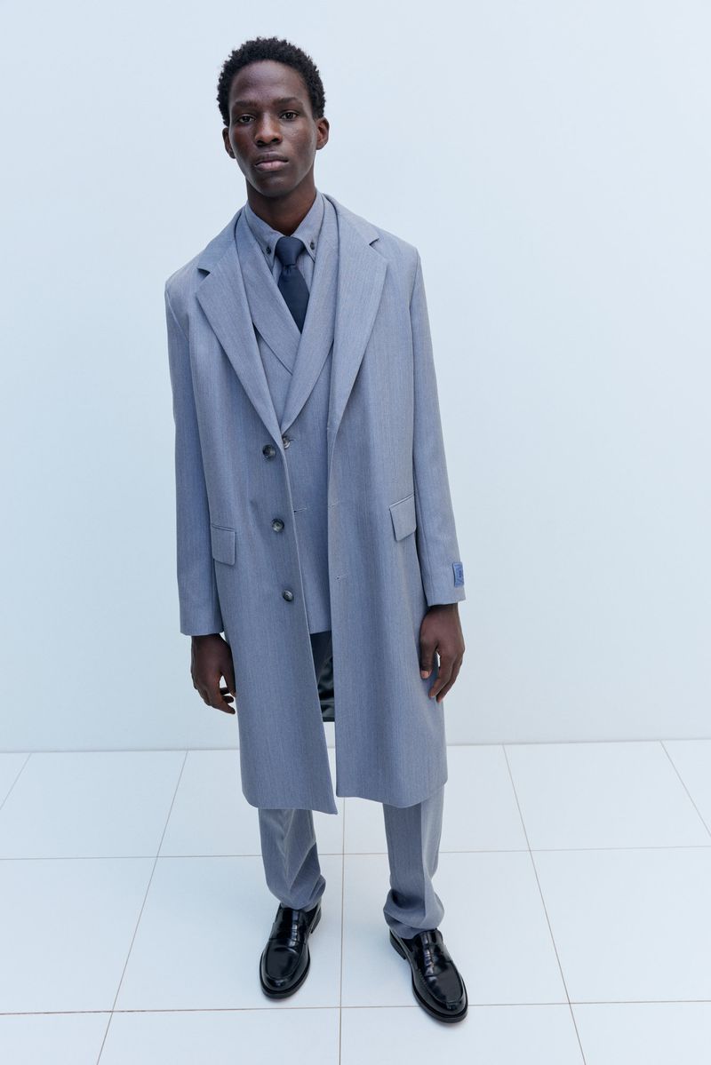 A tall, slender Black man wearing a grey three-piece suit and matching overcoat stands indoors against a light backdrop, fashion shoot.